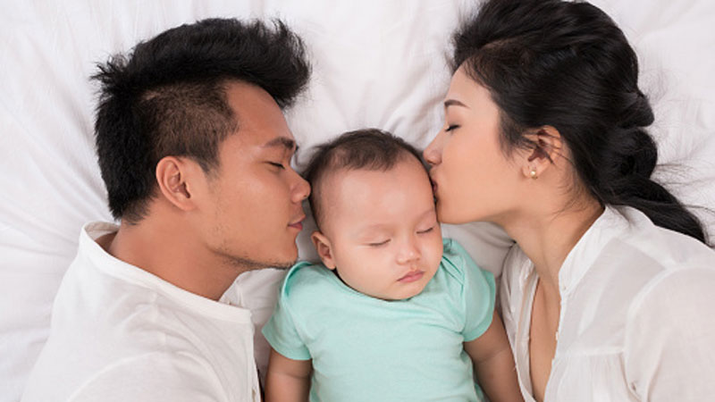 parental-leave-800x450 A baby with its eyes closed lying in a bed being kissed by its parents who are lying on both sides.